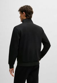 Zwarte jas met een hoge kraag, gemaakt van een soepele stof. Heeft elastische manchetten en een ruime pasvorm, gedragen met bijpassende zwarte broek.