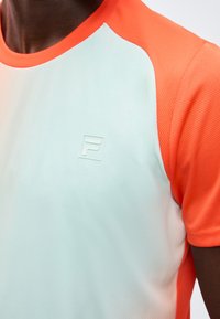 Κοντινό πλάνο ενός ατόμου που φοράει λευκό και πορτοκαλί αθλητικό κοντομάνικο με ένα μικρό ανάγλυφο λογότυπο "F" στο στήθος.