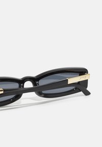 Sorte solbriller med en kraftig design, grå linser og et sideelement med gulldetaljer som er gravert med "pray". Har en elegant, glansfull finish.