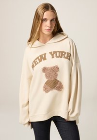 Haut à capuche couleur crème avec le texte "NEW YORK" et un graphique de nounours en peluche texturé. Matériau doux, coupe décontractée et manches longues avec ourlets côtelés.