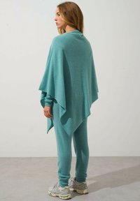 Poncho lavorato a maglia turchese con una vestibilità ampia e un orlo irregolare, abbinato a pantaloni aderenti coordinati e sneakers grigie. Texture complessiva liscia.