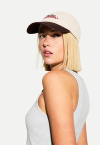 Beige kšiltovka s hnědým kšiltem, s červeným vyšitým textem. Modelka má na sobě šedý tílko, na kterém je vidět detailní profil.