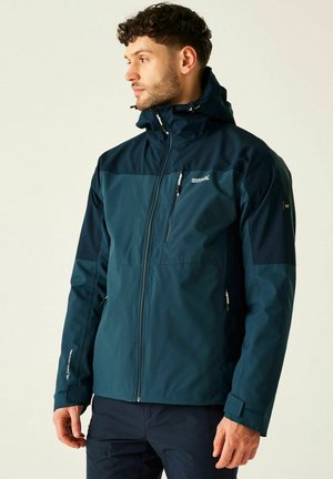 Mężczyzna w ciemnoturkusowej i granatowej kurtce outdoorowej na zamek z kapturem, stojący i patrzący w lewo na jednolitym tle.