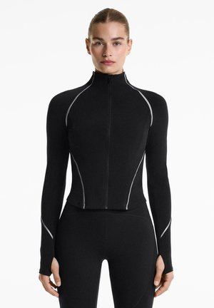 Giacca atletica nera aderente con maniche lunghe, colletto alto e dettagli riflettenti. Realizzata in un materiale elastico per comfort e libertà di movimento.