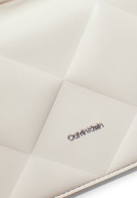Borsa a tracolla di Calvin Klein di colore crema, con una texture che presenta un motivo a quadri a diamante e un accento del logo argentato in basso a destra.