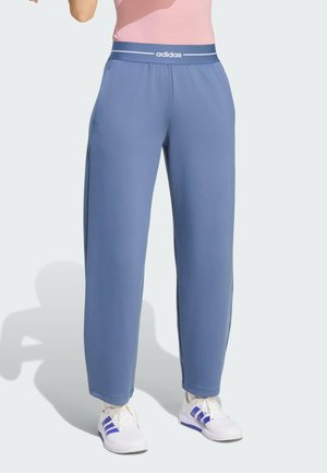 Osoba nosící modré tepláky Adidas s elastickým pasem a bílé tenisky s modrými pruhy na jednobarevném pozadí.