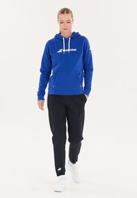 Blå hættetrøje med hvidt logo, forlomma og snor. Sorte sportsbukser med slim fit. Hvide sneakers med detaljer.