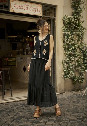 Un haut sans manches noir avec broderies florales et ourlet blanc, associé à une jupe noire à volants et des sandales à plateforme marron dans un cadre de pavés.