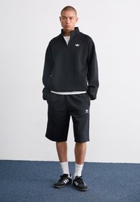 Sweat à fermeture éclair noir avec un logo blanc, associé à un short noir jusqu'aux genoux. Le modèle porte des chaussettes blanches et des baskets noires. Texture de tissu lisse.