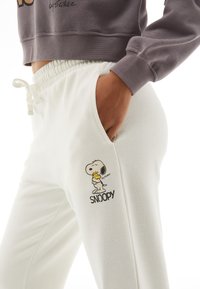 Pantalon de survêtement blanc en tissu doux, avec un graphique de Snoopy et du texte noir. Taille élastique avec cordon de serrage et poches latérales.