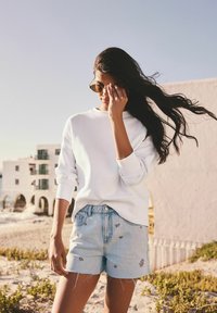 Sweatshirt blanc et short en denim bleu clair avec des détails de broderie florale. Des lunettes de soleil complètent la tenue décontractée de plage. Fond sablonneux.
