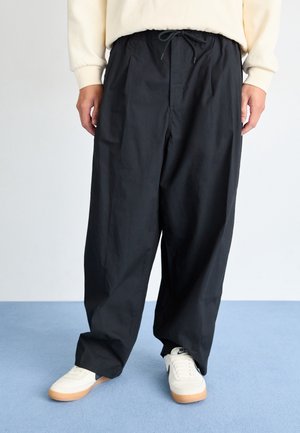 Person trägt lockere schwarze Hose mit Kordelzug an der Taille, cremefarbenen Pullover und weiße Sneaker mit Gummisohlen, steht auf blauem Teppich.