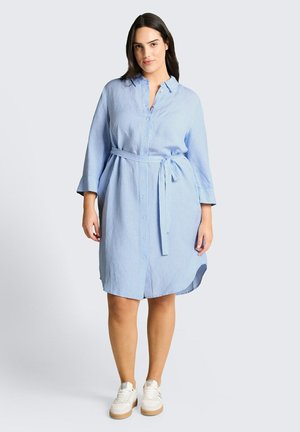 Kobieta ubrana w jasnoniebieską, długą koszulową sukienkę z paskiem w talii i białe trampki, stojąca na jednolitym tle.