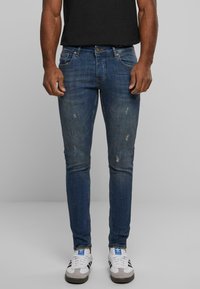 Jeans ajustados de color azul oscuro con áreas descoloridas y pequeños desgastes, hechos de tela de mezclilla. Combinados con zapatillas deportivas blancas y grises.
