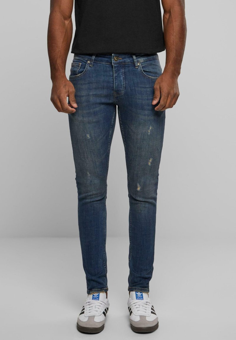 Jeans ajustados de color azul oscuro con áreas descoloridas y pequeños desgastes, hechos de tela de mezclilla. Combinados con zapatillas deportivas blancas y grises.