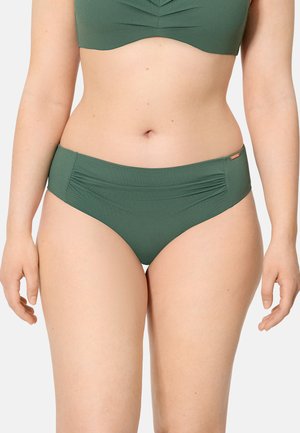 Mujer con bikini de tiro alto verde con fruncido sutil en los laterales y top de bikini a juego, de pie con los brazos relajados a los lados.