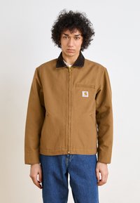 Chaqueta de lona marrón con un cuello oscuro en contraste, cierre delantero, bolsillos laterales y un bolsillo en el pecho con un parche de logo.