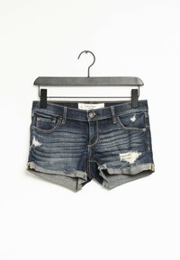 Shorts de mezclilla, azul oscuro con detalles desgastados, con dobladillo desflecado y puños enrollados, colgando de una percha de plástico negra.