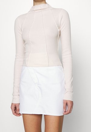 Langärmeliges, weich pinkfarbenes Oberteil mit hohem Ausschnitt und strukturierter Naht, kombiniert mit einem weißen Denimrock mit asymmetrischem Design.