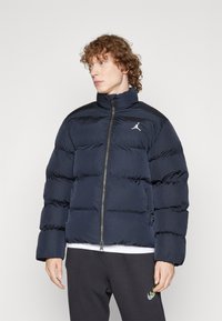 Mornarska puff jakna z visokim ovratnikom, prešito zasnovo, sprednjim zadrgo in majhnim belim logotipom na levi strani. Ima stranske žep