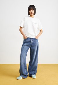 Hvid bomulds t-shirt med et lille grafik, kombineret med løstsiddende blå denim jeans og grønne sko med hvide såler på et gult gulv.