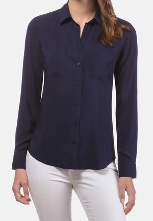 Kobieta ubrana w granatową, zapinaną na guziki koszulę z długim rękawem i dwoma kieszeniami na piersi, połączoną z białymi spodniami, na jednolitym tle.