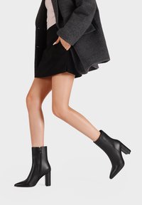 Stivali ankle in pelle nera con punta affusolata, chiusura con zip e tacco spesso, esposti accanto a un cappotto grigio e a una gonna nera.