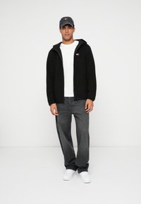 Musta fleece-takki hupulla, valkoinen t-paita, harmaat farkut ja valkoiset lenkkarit. Lippis on logolla ja tummanharmaasta kankaasta.