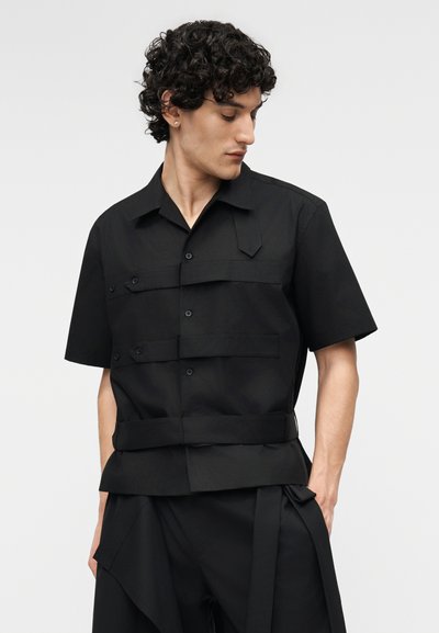 Νεαρός άνδρας με σγουρά μαλλιά, φοράει μαύρο πουκάμισο με κοντά μανίκια με κουμπιά και πολλές τσέπες, και αντίστοιχο μαύρο παντελόνι, κοιτάζει προς τα αριστερά.