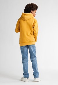 Felpa gialla con cappuccio e tasca a marsupio, abbinata a jeans azzurri chiari e sneakers bianche, caratterizzata da un design semplice e informale.
