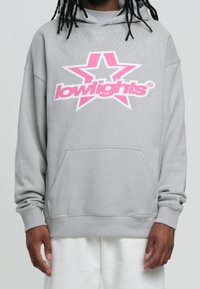 Sudadera gris con un gran gráfico de estrella rosa y blanca en el centro, con el texto "lowlights", bolsillo frontal tipo canguro y puños de canalé.