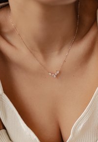 Naszyjnik w kolorze różowego złota z małymi, przejrzystymi kamieniami w kształcie łzy ułożonymi w kształt litery V. Cienki łańcuszek o delikatnym, teksturowanym wzorze.
