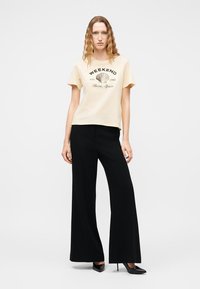 Femme aux cheveux ondulés portant un t-shirt beige "Weekend Ibiza Spain", un pantalon large noir et des talons hauts noirs, debout devant un fond blanc.