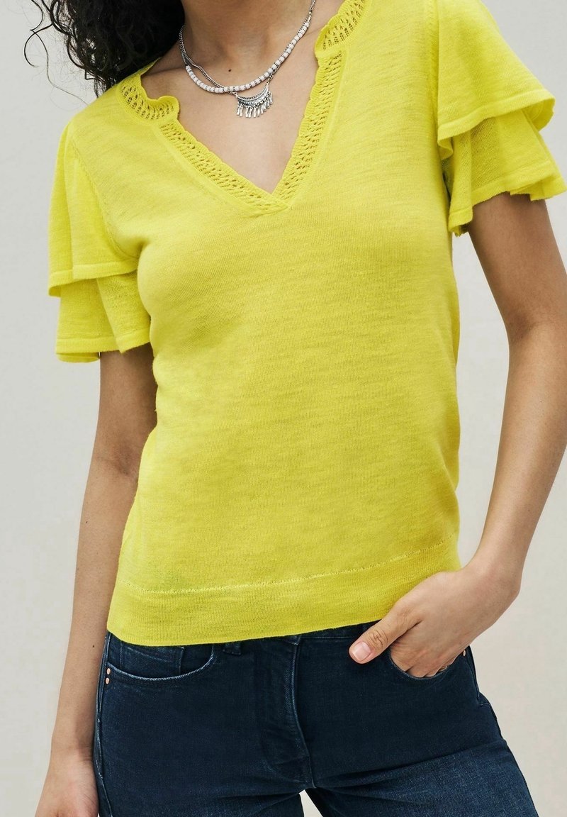 Femme portant un haut en maille jaune à manches courtes avec des manches volantées et un col en V festonné, associé à un jean bleu foncé et des colliers argentés.