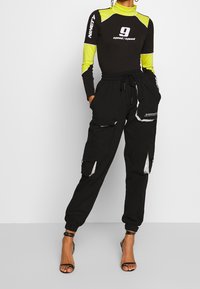 Zwart longsleeve bodysuit met gele accenten en grafische print, gecombineerd met zwarte cargo broek met reflecterende details en een elastische tailleband.