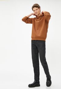 Bruine hoodie met een trekkoord en cuffed mouwen, gecombineerd met zwarte slim-fit jeans en zwarte enkellaarsjes, tegen een witte achtergrond.
