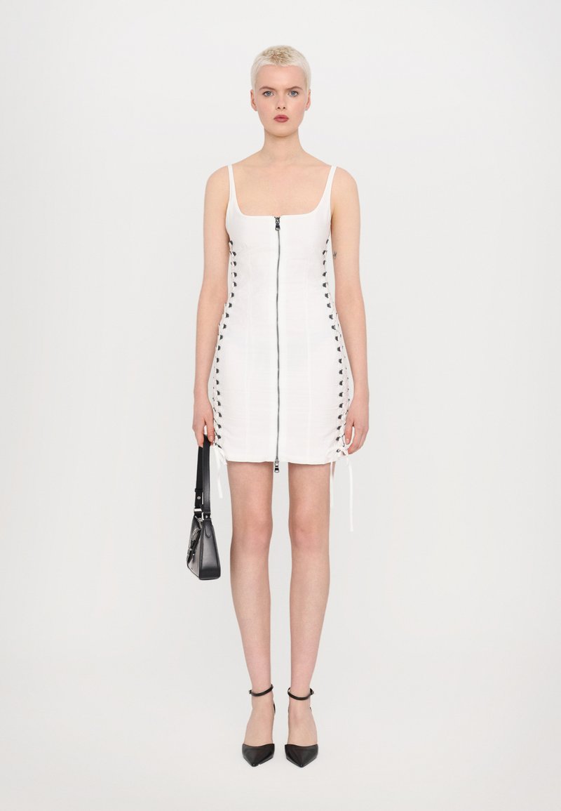 Witte mouwloze jurk met rits aan de voorkant, details met zijsnoeringen en trekkoordjes. Het model houdt een kleine zwarte handtas vast en draagt zwarte puntige hakken.