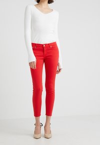 Mujer vistiendo unos pantalones vaqueros ajustados rojos, una blusa blanca de manga larga de canalé con agujeros para los pulgares y unos zapatos de tacón de punta beige con correas negras en los tobillos.