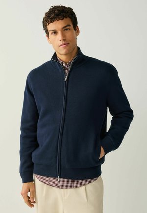 Navy zip-up jacket with ribbed cuffs and hem, made of soft fabric; layered over a red and white checked shirt.
