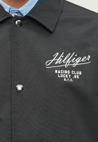 Μαύρο μπουφάν με υφασμάτινη επιφάνεια, που διαθέτει κλείσιμο με κουμπί, κομψό γιακά και λευκή κεντημένη επιγραφή: "Hilfiger Racing Club Lucky .85 N.Y.C."