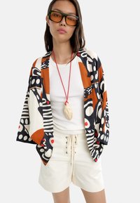 Dziennikarka kimono z nadrukiem w wielokolorowe wzory abstrakcyjne, zestawiona z białą bluzką o prążkowanej fakturze i beżowymi szortami z wiązaniem z przodu.