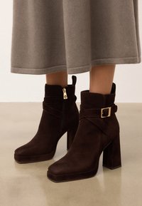 Brązowe zamszowe botki na kostkę z kwadratowym noskiem, masywnym obcasem, zapięciem na zamek błyskawiczny oraz z klamrą wokół kostki. Płaska podeszwa.