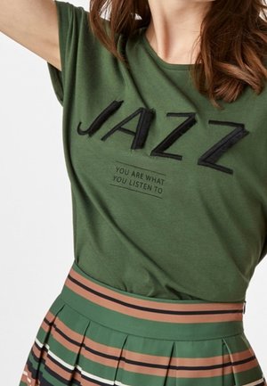 Vrouw draagt een groen T-shirt met de tekst "JAZZ" en "YOU ARE WHAT YOU LISTEN TO", gecombineerd met een geplooide rok met groene, bruine en zwarte strepen.