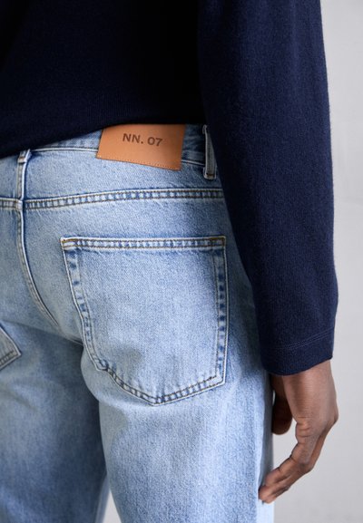 Lyseblå jeans med teksturert finish, med et brunt lærmerke merket "NN. 07" på baklinning og synlige sømdetaljer.