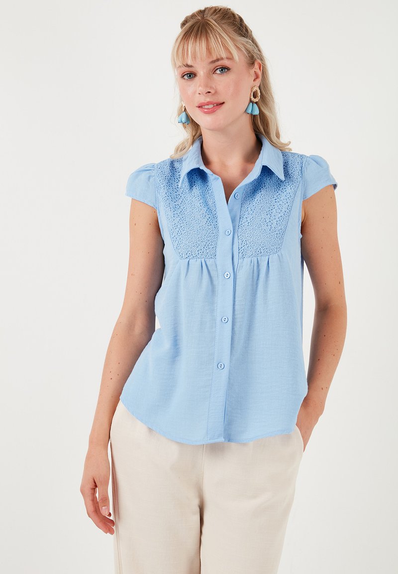 Blusa azzurra chiara a maniche corte con bottoni sul davanti e dettagli in pizzo sulle spalle, realizzata in un tessuto morbido e testurizzato.