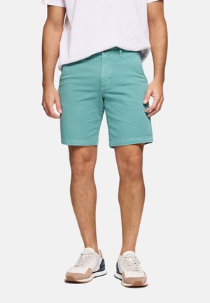 Hombre con pantalones cortos color aguamarina hasta la rodilla, zapatillas blancas y camiseta blanca, de pie con los brazos relajados a los lados sobre fondo blanco.