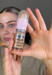 Maybelline Perfector 4-i-1 Glow makeup i en transparent flaska med ett rosa lock. Etiketten visar nyansen "1.5 Light Medium" och produktens egenskaper.