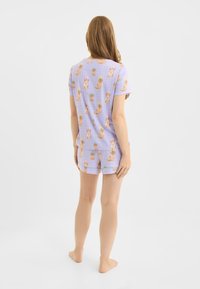 Kort pyjama setje gemaakt van lichtgewicht stof. Het heeft een lavendelkleurige achtergrond met een speels hondenpatroon en een rasterontwerp.