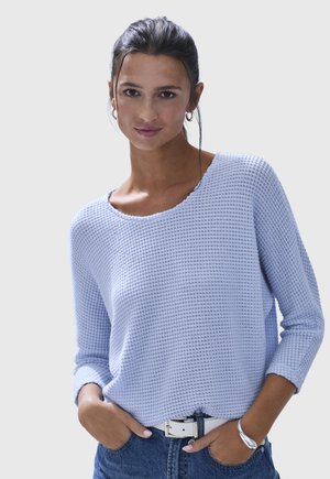 Junge Frau mit zurückgebundenem dunklem Haar, die einen strukturierten hellblauen Pullover, blaue Jeans, weißen Gürtel und ein silbernes Armband trägt, die Hände in den Taschen.