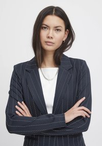 Marineblauer, gestreifter Blazer mit tailliertem Design, grauer Naht, Reverskragen und slim fit. Über einem weißen Oberteil getragen.